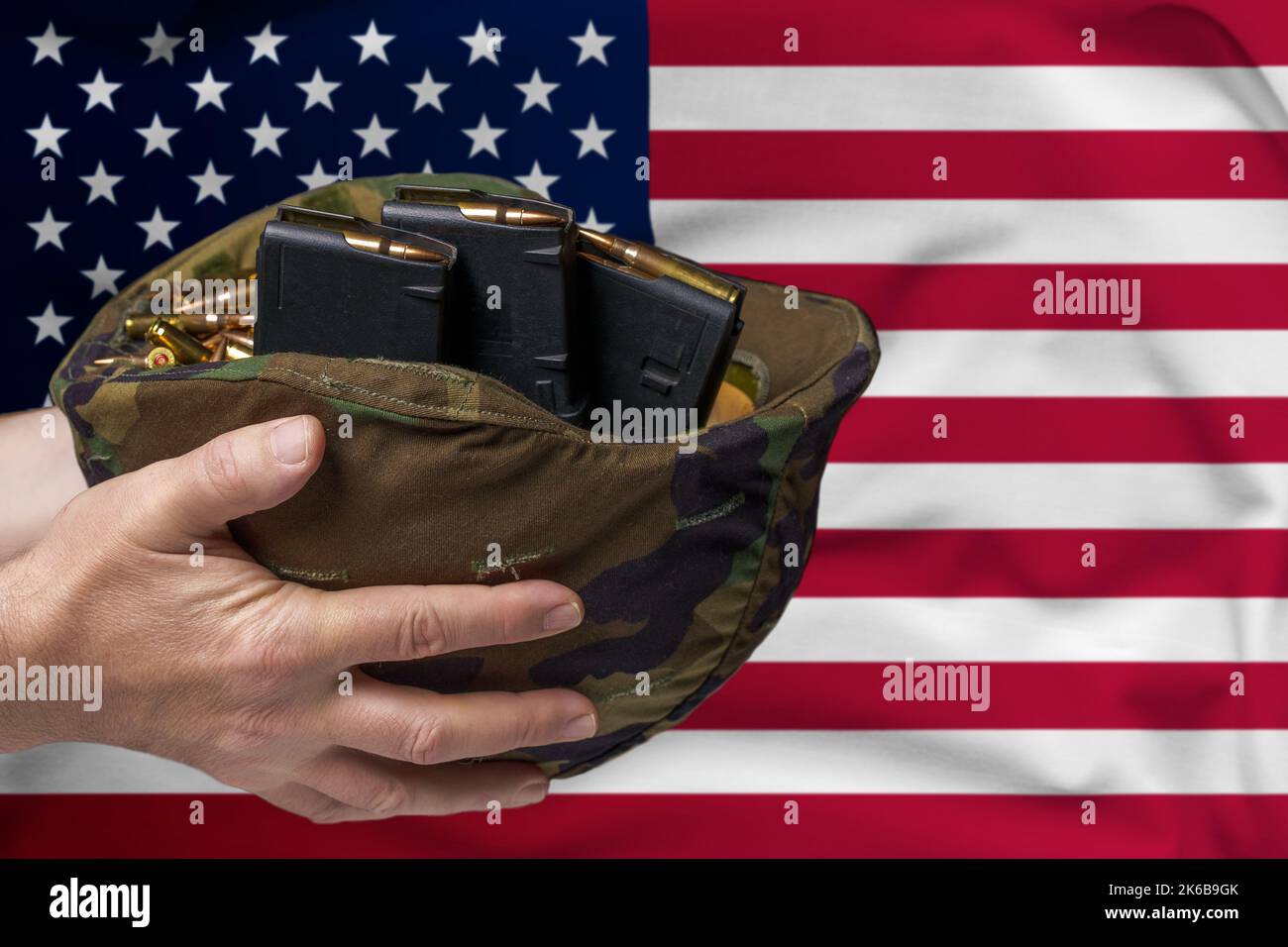 Ein Militärhelm mit Patronen und Zeitschriften für ein Gewehr in den Händen eines Mannes vor dem Hintergrund der Flagge der USA. Das Konzept des Verkaufs Stockfoto