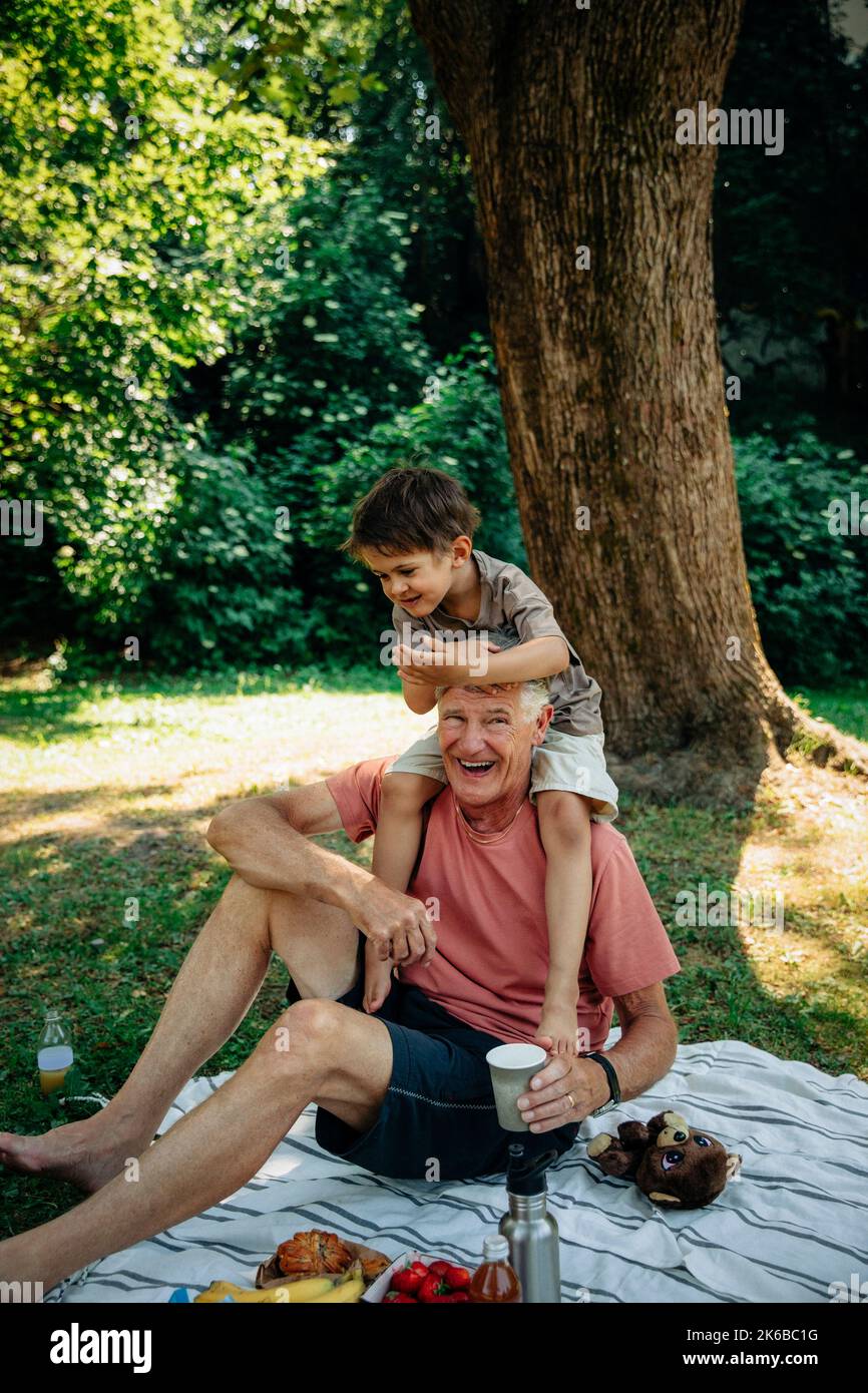 Großvater trägt Enkel auf der Schulter, während er auf einer Picknickdecke im Park sitzt Stockfoto