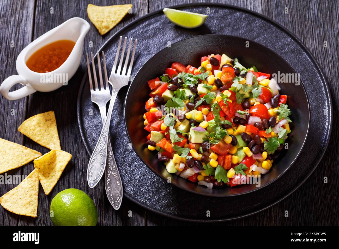 Cowboy-Kaviar, texas-Kaviar mit schwarzer Bohne, Tomaten, Avocado, roter Paprika, Mais, Koriander in schwarzer Schale auf dunklem Holztisch mit Nachos und d Stockfoto