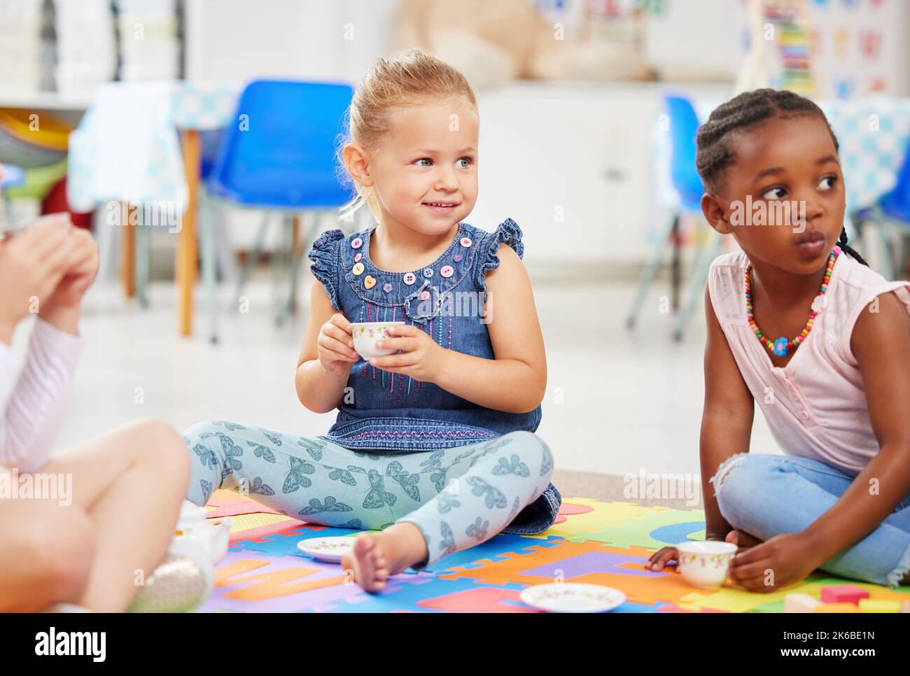 Kleine Mädchen, die eine Teeparty machen, während sie auf einer bunten Matte auf dem Boden in der Vorschule oder im Kindergarten spielen. Fröhliche, vielfältige Kinder, die Spaß haben Stockfoto