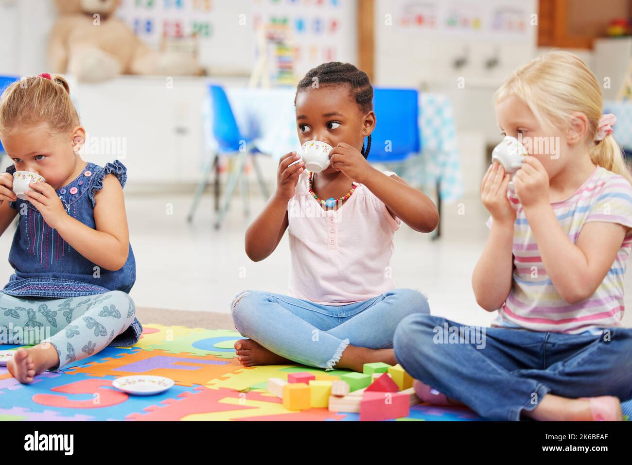 Eine Gruppe verschiedener kleiner Mädchen, die eine Teeparty machen, während sie auf einer bunten Matte auf dem Boden in der Vorschule oder im Kindergarten spielen. Kinder mit gemischter Rasse Stockfoto