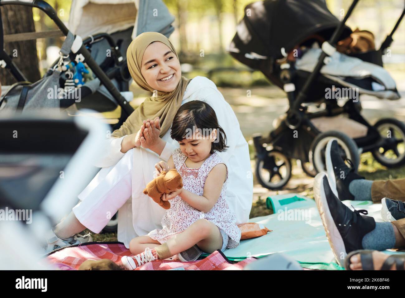 Lächelnde Frau im Hijab, die wegschaut, während sie mit der Tochter sitzt, die einen Teddybären hält Stockfoto