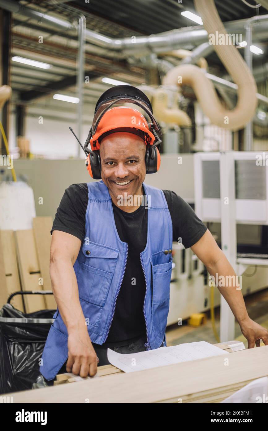 Porträt eines glücklichen Zimmermanns mit Hardhut und Ohrenschützern, der im Lager arbeitet Stockfoto