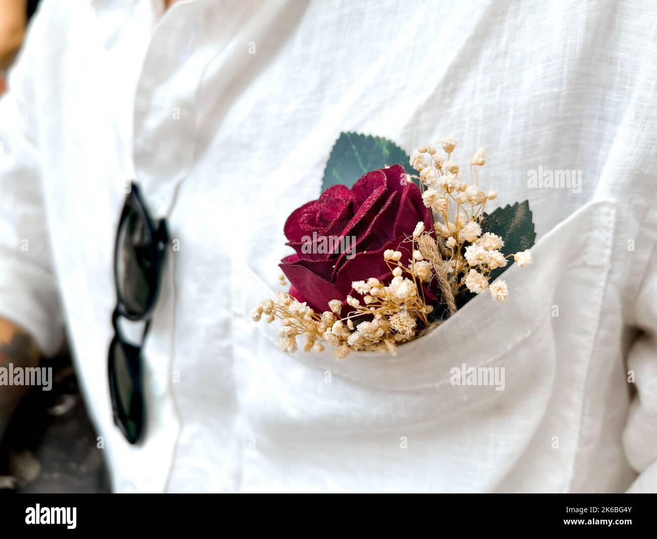 Nahaufnahme der Boutonniere in der Tasche eines weißen Leinenhemdes mit Sonnenbrillen. Selektiver Fokus auf rote Rose. Stockfoto