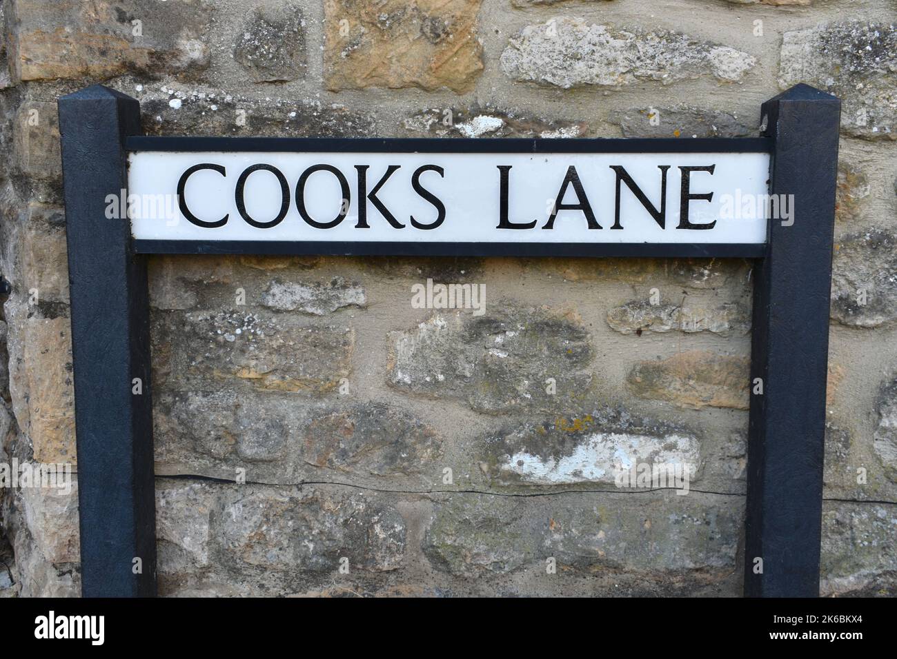 Nahaufnahme des Straßenschilds Cooks Lane auf einem alten Steinmauer Hintergrund. Das Vintage-Textschild ist schwarz auf weißem Metall-Hintergrund geschrieben. Stockfoto