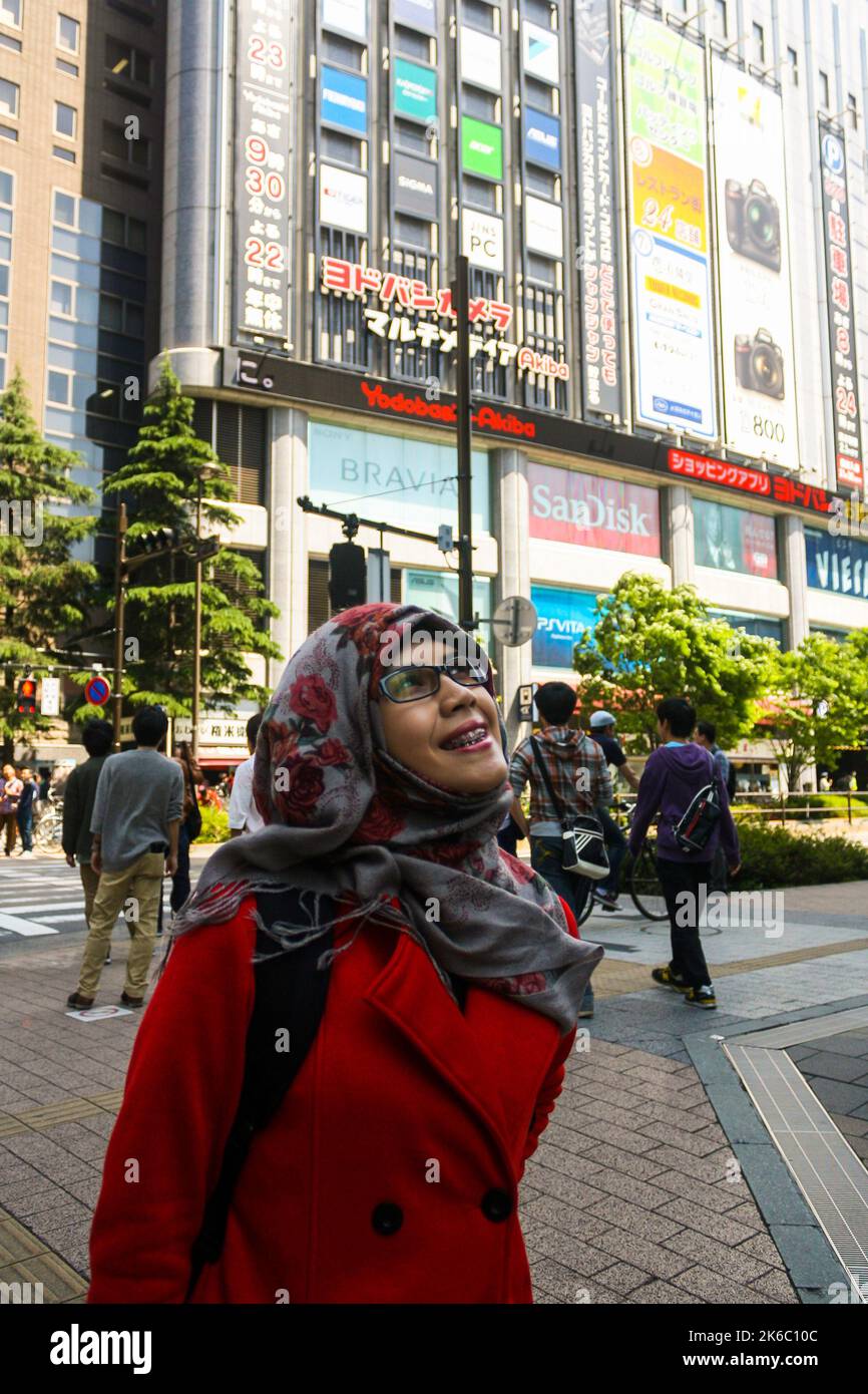 Portait einer jungen muslimischen Frau, die mit dem Yodobashi-Akiba-Gebäude lächelt, und Menschenmenge, die in Akihabara spazieren und einen klaren blauen Himmelshintergrund haben. Stockfoto