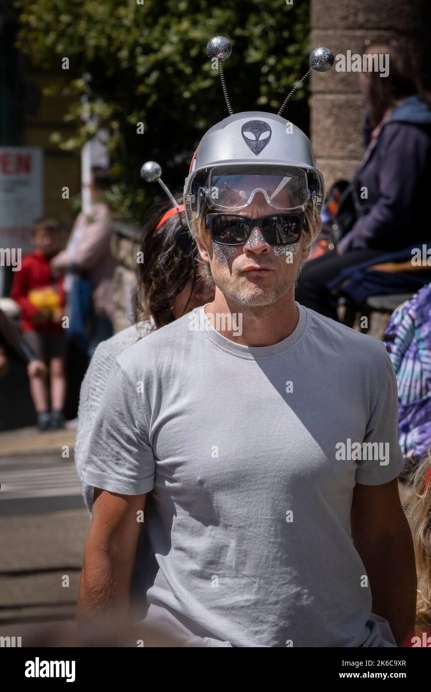 Ein Mann mit einem bemalten Gesicht, der eine Sonnenbrille und einen silbernen Sicherheitshelm trägt, der als Alien dekoriert ist. Stockfoto