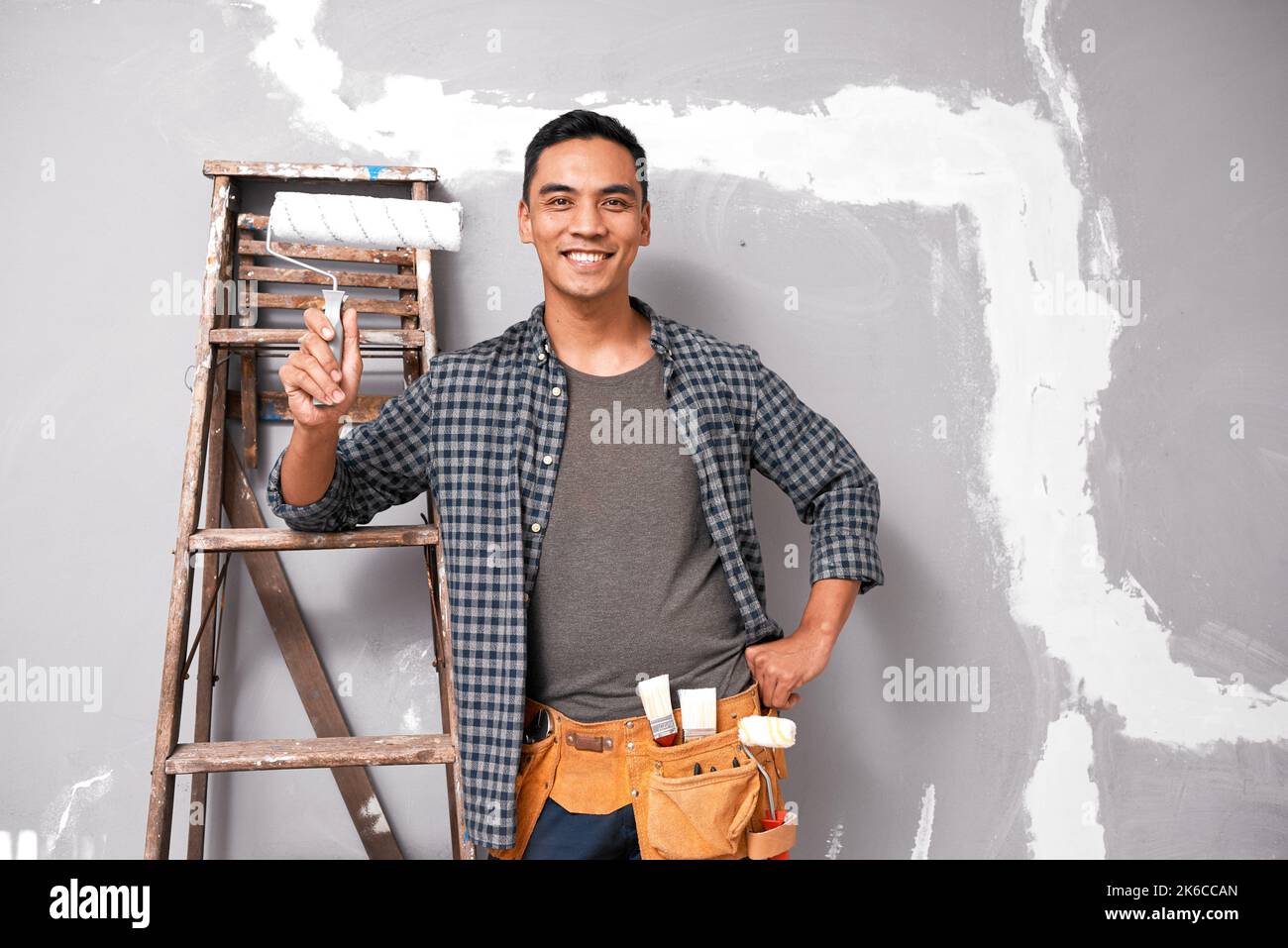 Ein attraktiver asiatischer Mann lächelt und posiert mit Malgeräten gegen die Wand Stockfoto