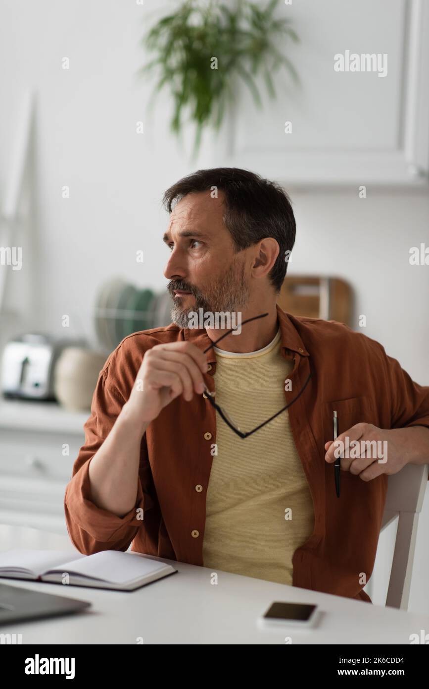 Nachdenklicher Mann, der eine Brille hält und in die Nähe eines leeren Notizbuchs in der Küche schaut, Stockbild Stockfoto