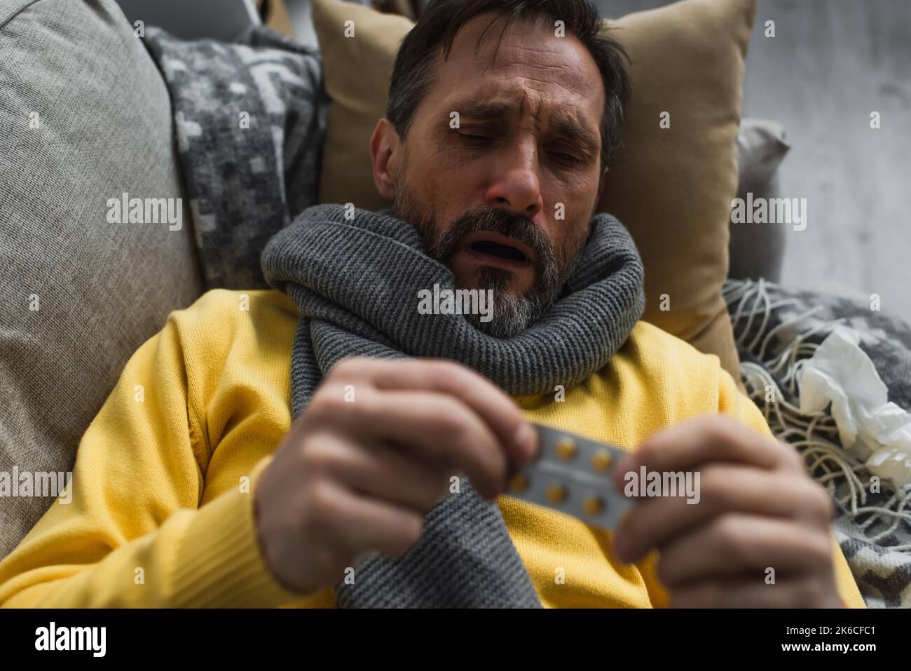 Kranker Mann in warmem Schal mit verschwommenen Pillen, während er auf dem Sofa lag, Stockbild Stockfoto