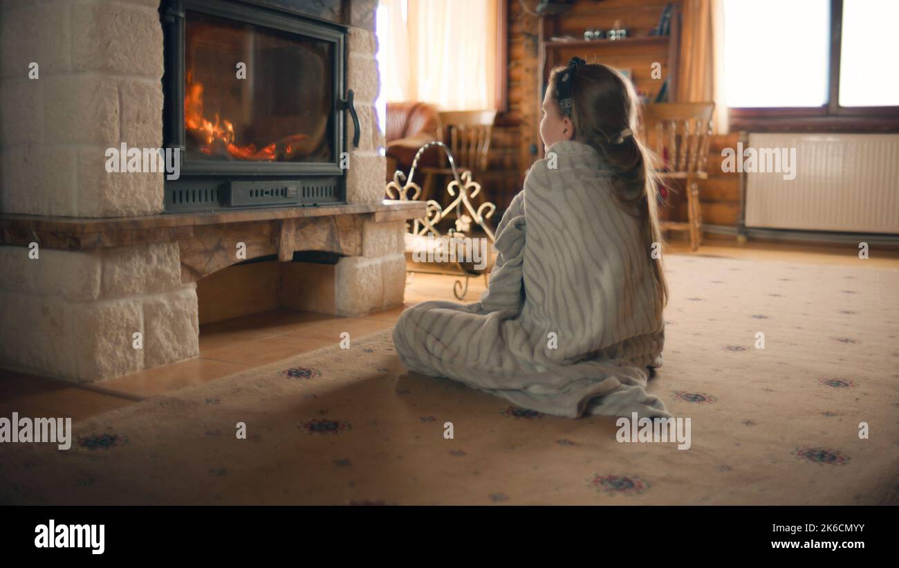 Junges Mädchen, das am Kamin auf einem milden Teppich sitzt, am Feuer zuschaut und sich an kalten Tagen mit Deckchen erwärmt, entspannt und Freizeit zu Hause verbringt Stockfoto