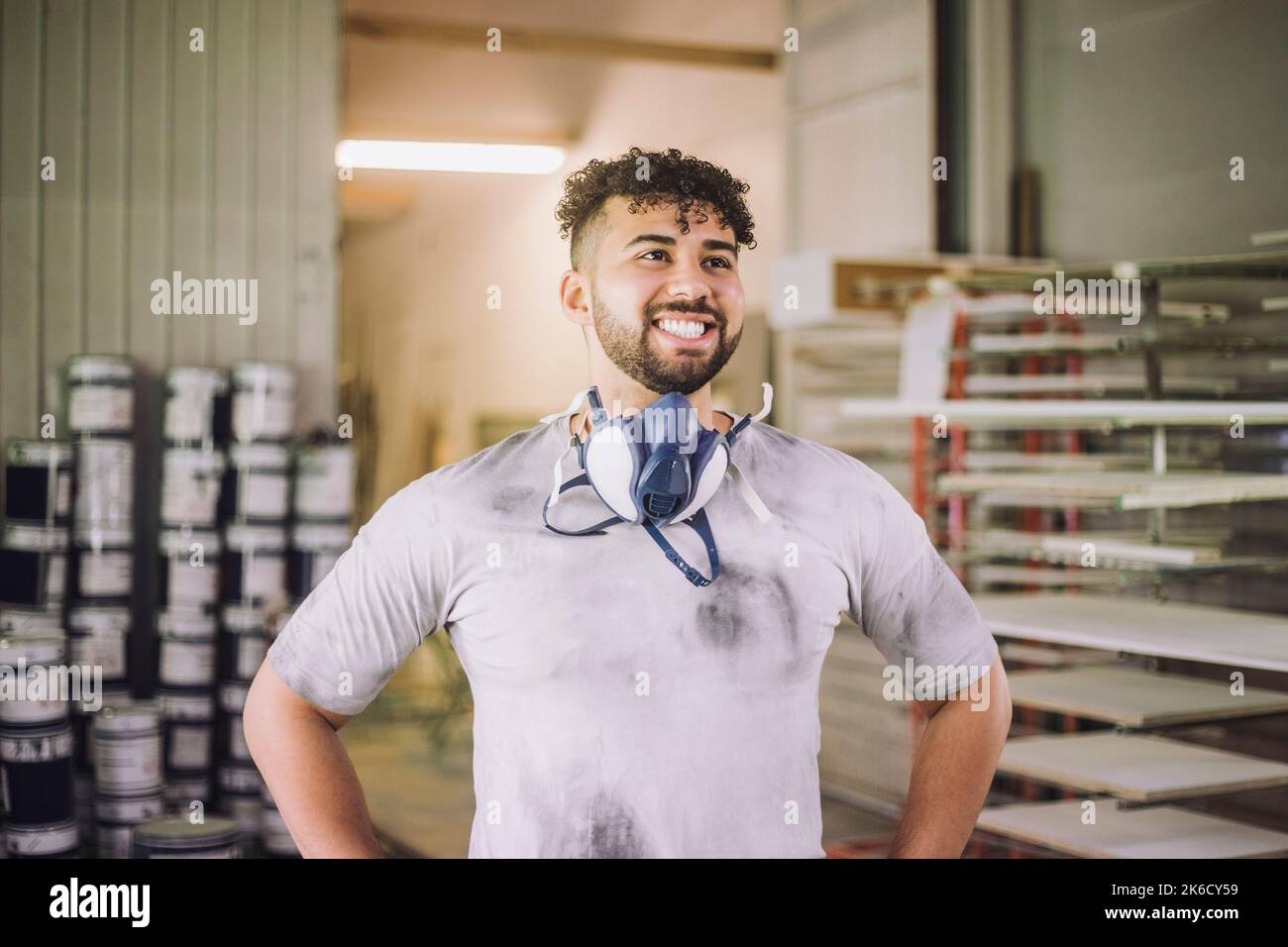 Fröhlicher junger Maler, der in der Werkstatt mit den Händen auf den Hüften steht Stockfoto