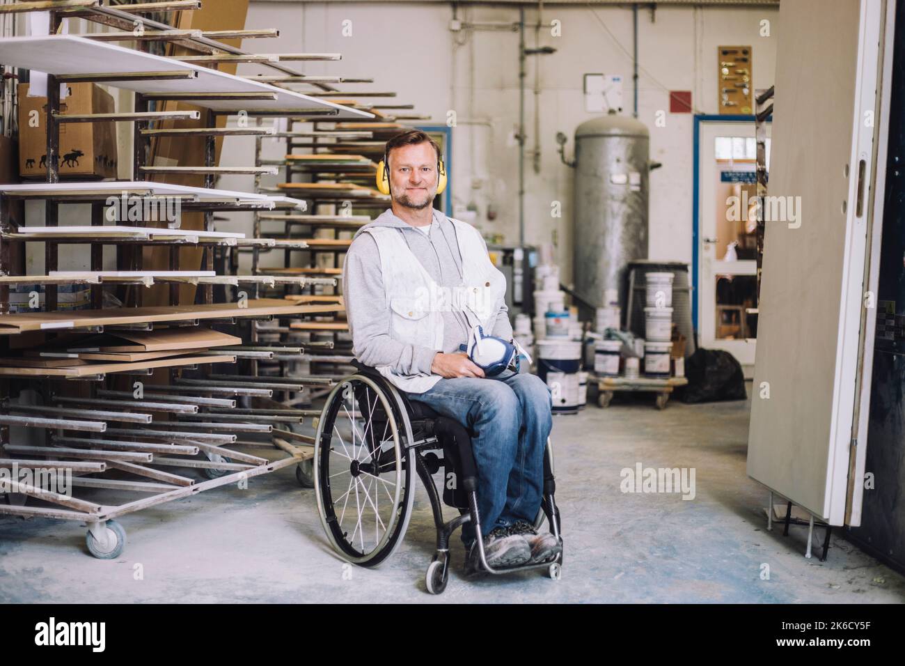 Lächelnder Maler mit Ohrenschützern, der in der Werkstatt auf dem Rollstuhl sitzt Stockfoto