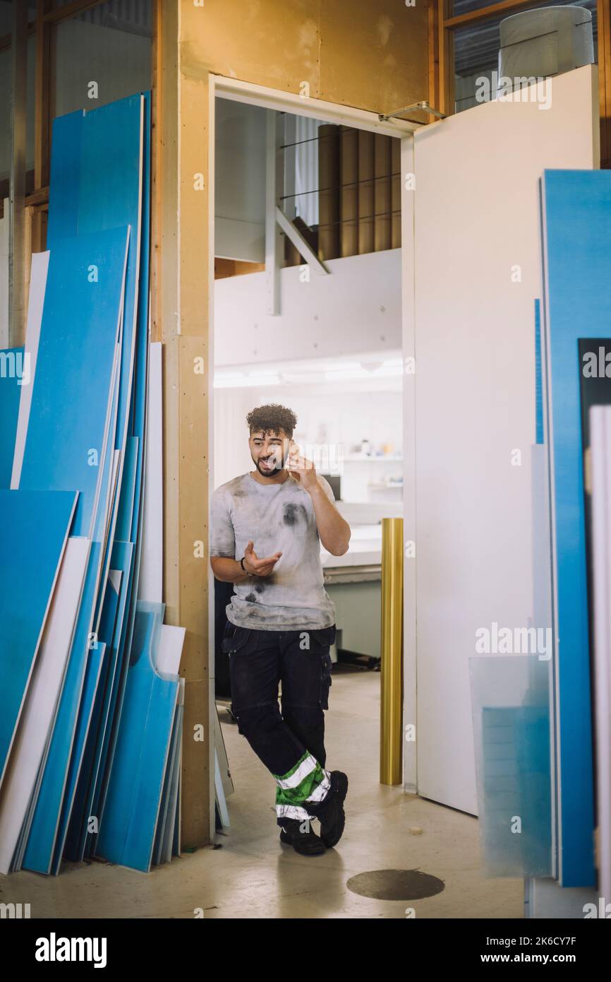Die ganze Länge des jungen männlichen Zimmermanns, der auf dem Smartphone spricht, während er vor der Tür der Werkstatt steht Stockfoto