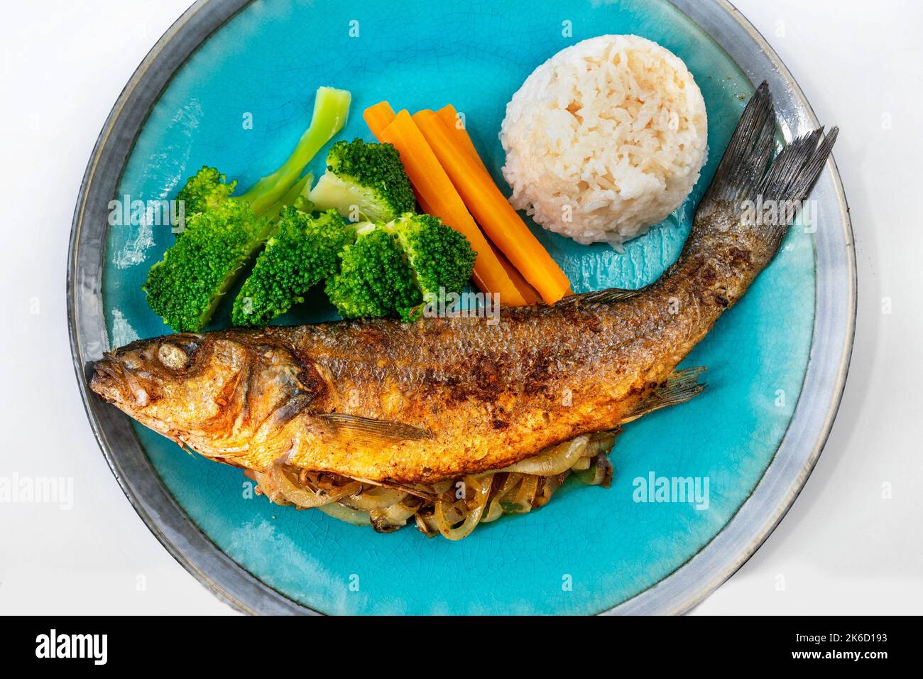 Gebratene Forelle gefüllt mit Zwiebeln, Gemüsegarnitur (Brokkoli und Karotte) und Reis auf blauem Teller, Nahaufnahme. Stockfoto