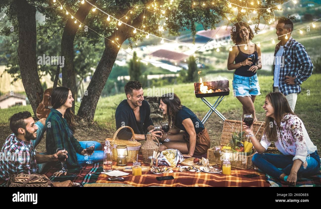 Fröhliche Freunde, die nach Sonnenuntergang im Weinberg Spaß haben - Jugendliche zelten tausendjährig beim Open Air Picknick unter Glühbirnen - Jugendfreundschaftskonzept wi Stockfoto