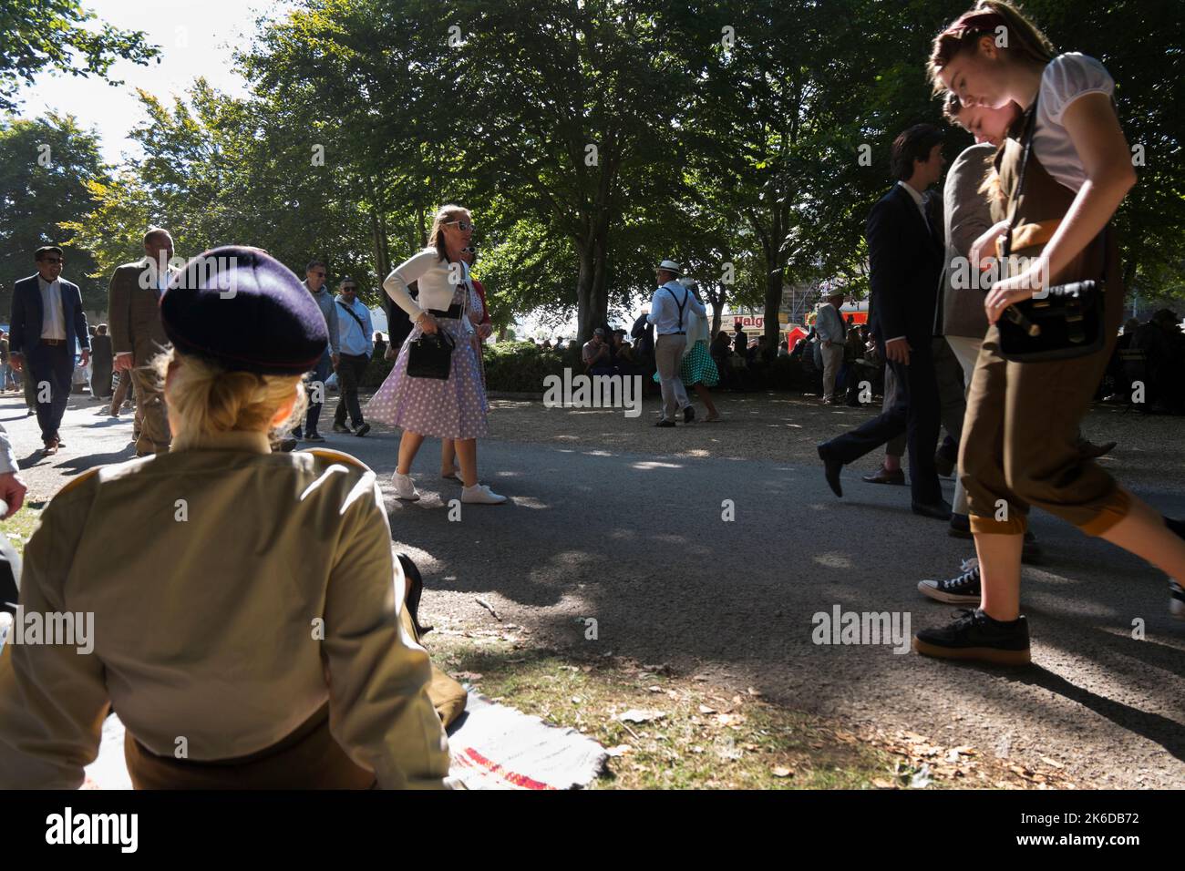 Menschen in Vintage-Kleidung, die an einer Frau in ATS-Uniform vorbeigehen, die auf dem Gras sitzt, BARC Revival Meeting, Goodwood-Rennstrecke, Chichester, Großbritannien Stockfoto