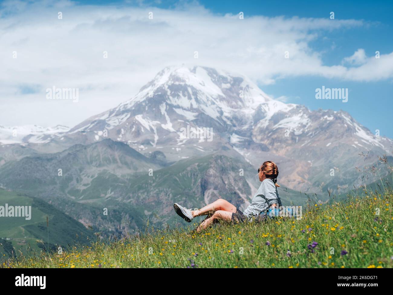 Backpacker Frau sitzt auf einem grünen Grashügel und genießt verschneite Hänge des Kazbek 5054m Berg mit einem Rucksack, während sie auf grünem Gras hil zu Fuß Stockfoto