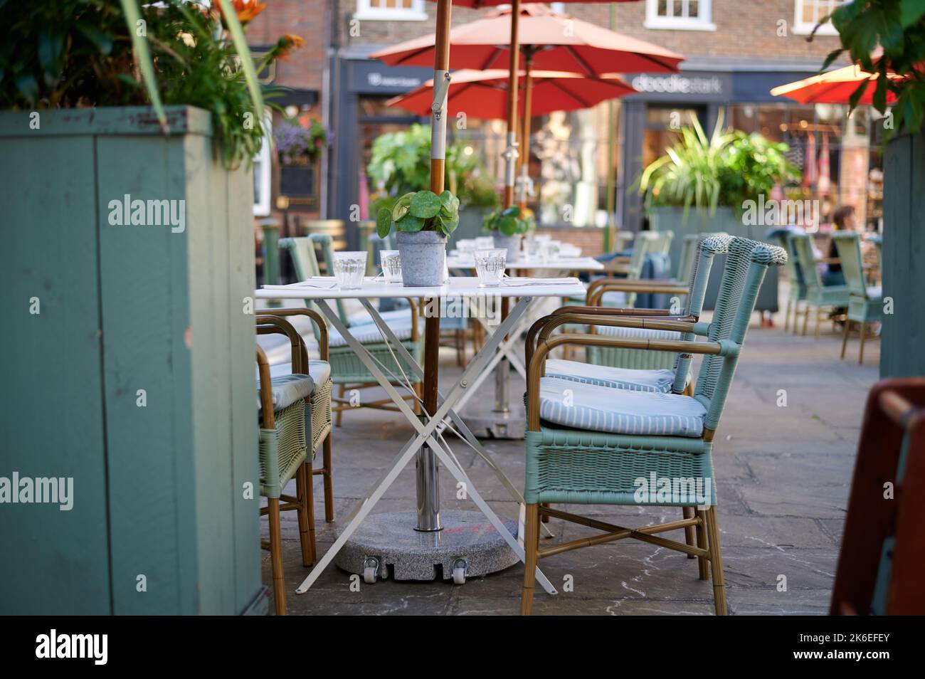 Leere Klapptische und großer Sonnenschirm / Sonnenschirm im Café mit Pflanzen, Glasküssen und Korbstühlen Stockfoto