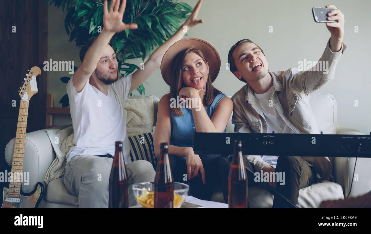 Verspielte junge Leute aus der Musikband machen Selfie mit dem Smartphone, posieren und machen lustige Gesichter, lachen und umarmen. Musikinstrumente Keyboard und Gitarre sind sichtbar. Stockfoto