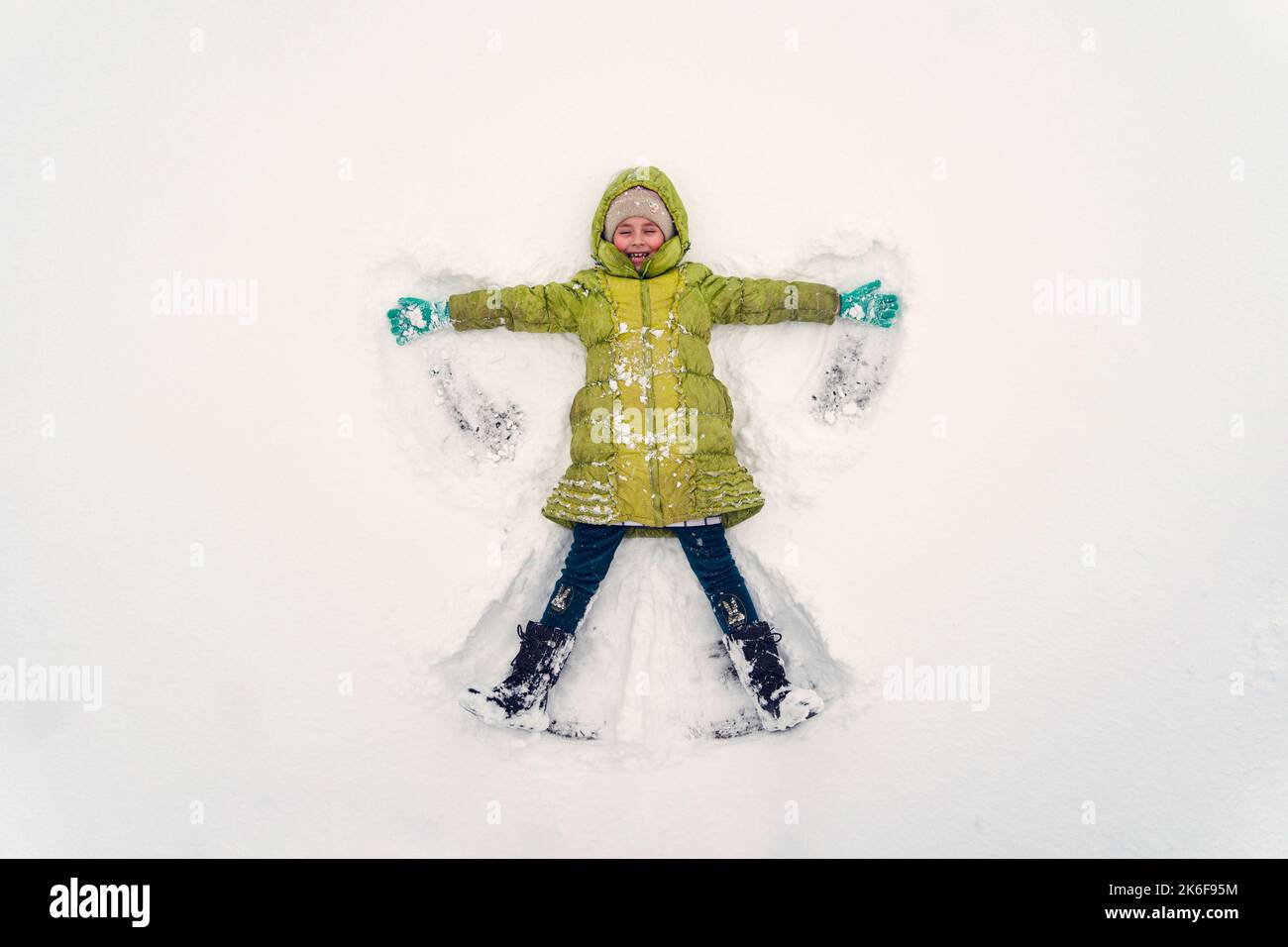 Aktive Kinder in einer grünen Jacke im Freien am Weihnachtstag. Kind macht Schnee Engel am sonnigen Wintermorgen. Kinder im Winter Spaß im Freien. familienweihnachtszeit vac Stockfoto