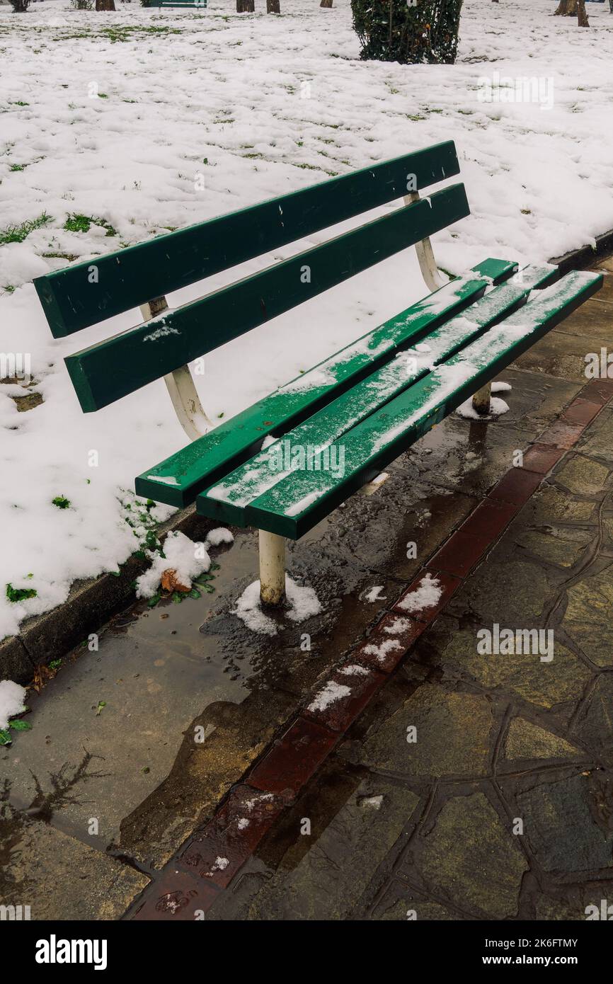 Tagesansicht einer leeren grünen Holzbank auf einem teilweise schneebedeckten Parkgelände. Stockfoto
