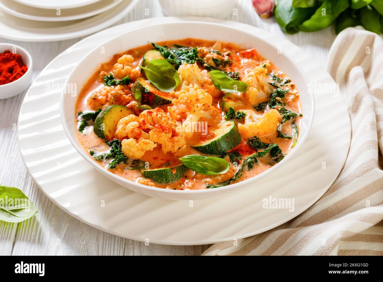 Cremige Tomatensuppe mit gemahlenem Huhn, Blumenkohl, Zucchini, Grünkohl, Basilikum und Gewürzen in einer weißen Schüssel auf einem weißen Holztisch, Blick auf die Landschaft Stockfoto