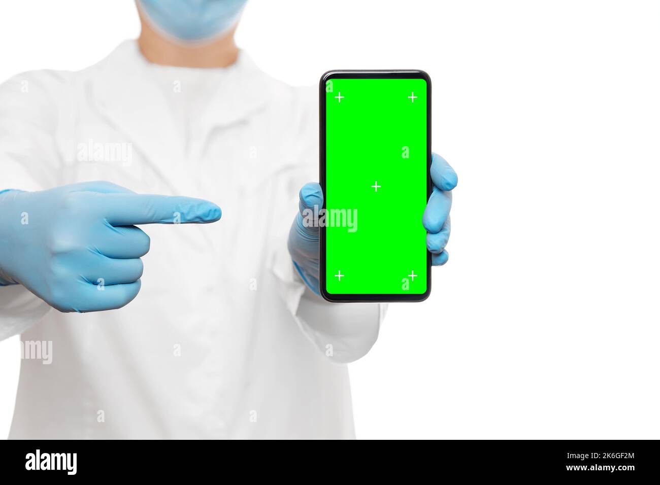 Ausgeschnittene Aufnahme eines Arztes in einer blauen medizinischen Maske und Handschuhen, die das Smartphone mit Greenscreen und Trackingpunkten halten und mit dem Finger darauf zeigen. Isoliert ein Stockfoto