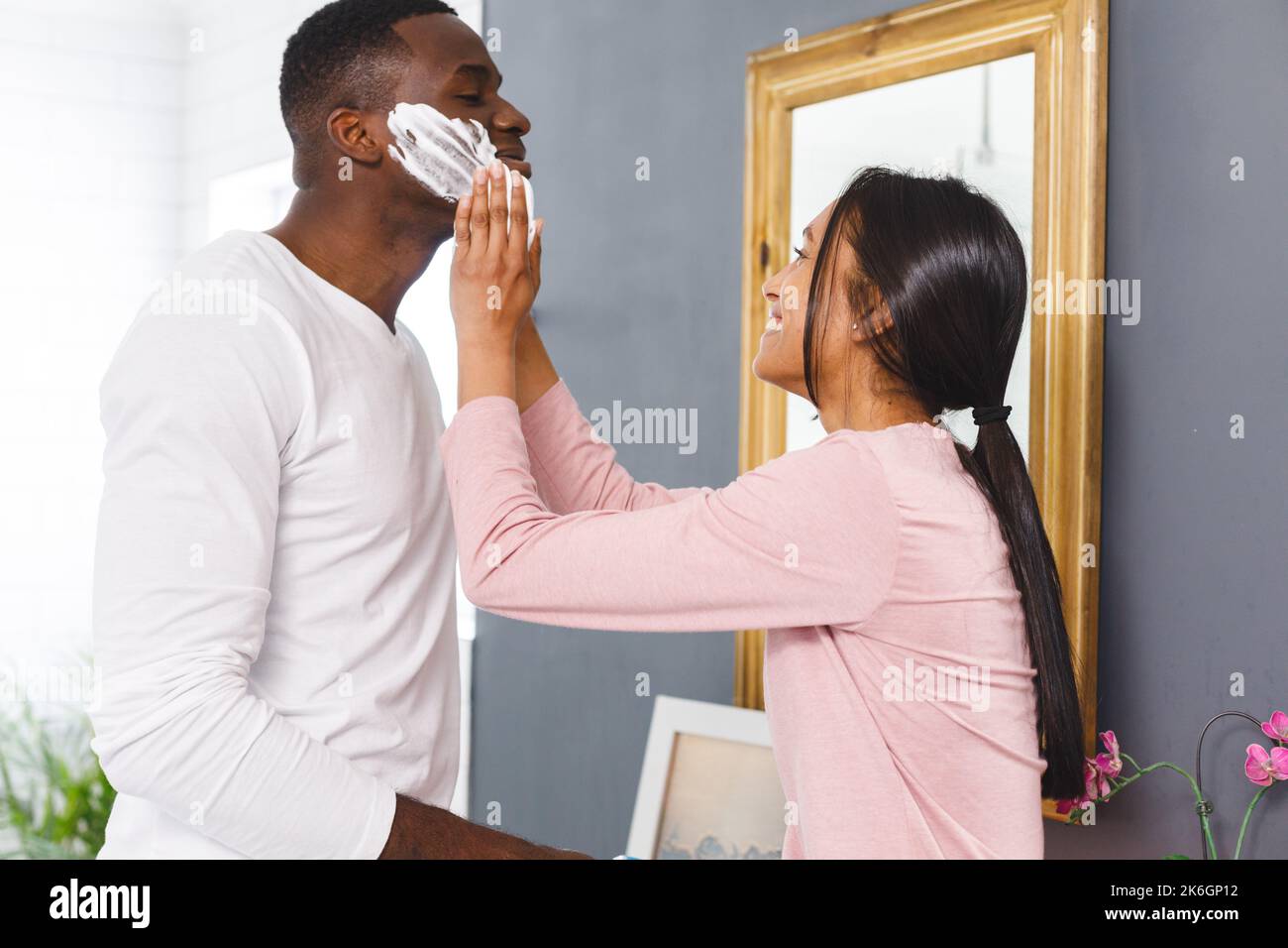 Glückliches, vielseitiges Paar im Badezimmer, Frau, die Rasiercreme auf das Gesicht eines Mannes legt, lächelt Stockfoto