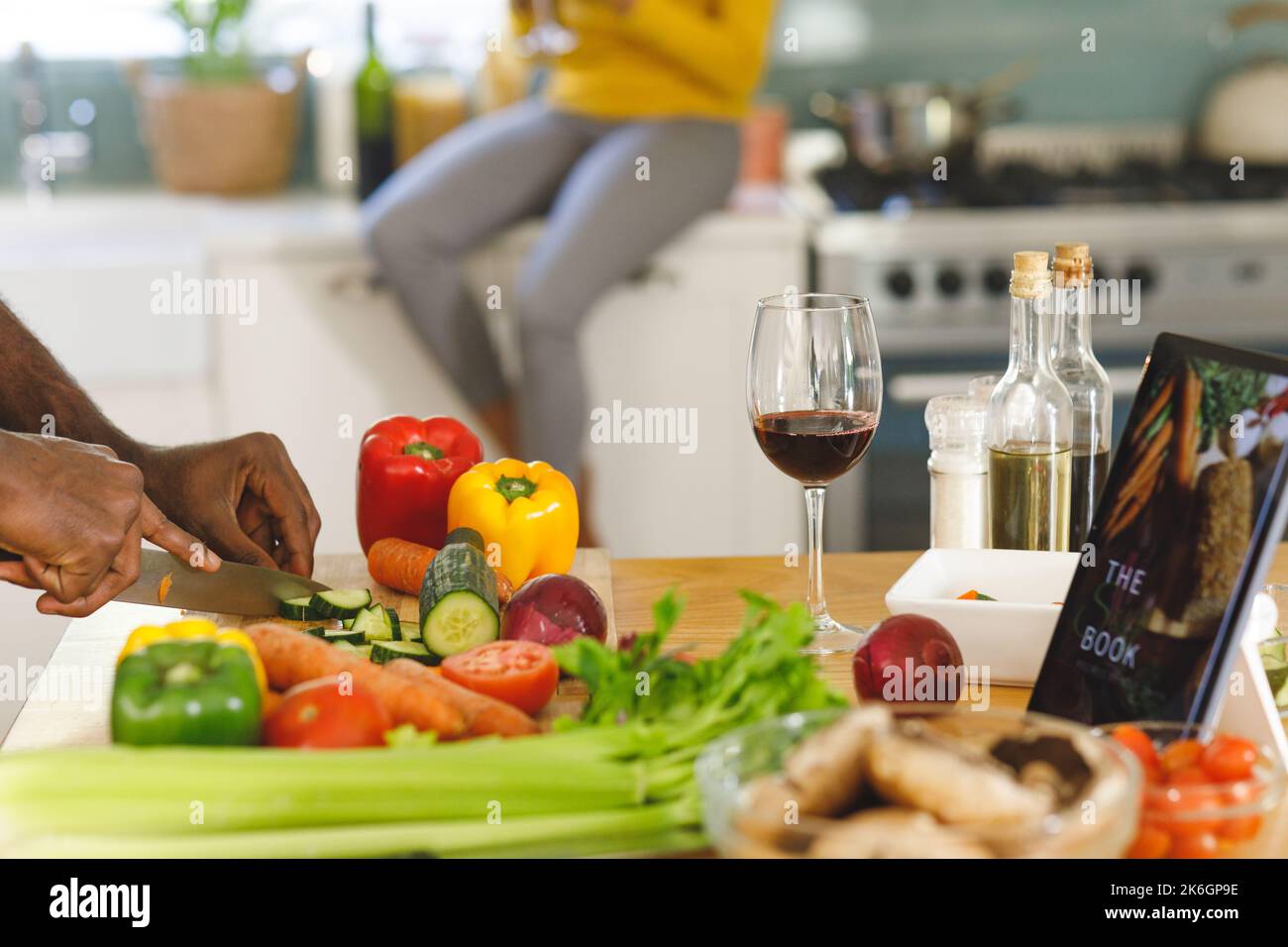 Hände eines afroamerikanischen Mannes mit Rezept auf einem Tablet, der in der Küche Lebensmittel zubereitet, Partner im Hintergrund Stockfoto