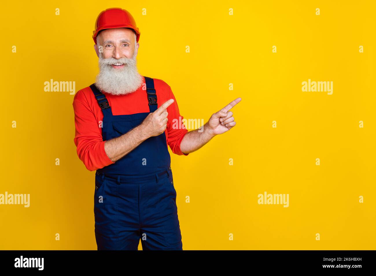 Foto von lustigen fröhlichen Alter Mann Arbeitskleidung insgesamt roten Hut zeigt Finger leeren Raum isoliert gelben Hintergrund Stockfoto