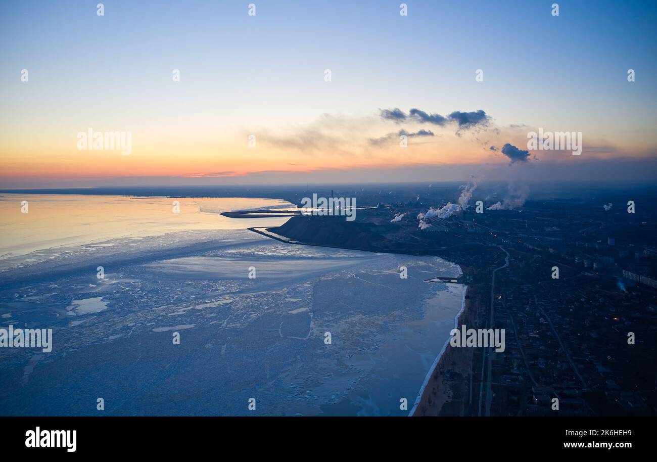 Luftaufnahme eines Industriewerks an der Küste eines gefrorenen Asowschen Meeres in Mariupol. Draufsicht auf die Fabrik, Rauch steigt aus den Schornsteinen. Stadt in der Nähe von Eis Stockfoto