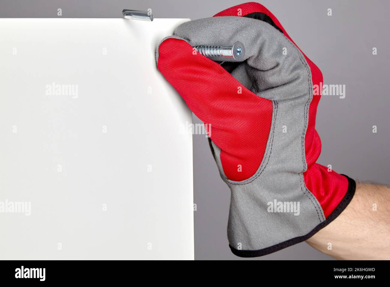 Eine Hand in einem roten Schutzhandschuh führt eine Bestätigung in ein Loch in einem Möbelschild ein. Montage und Reparatur von Möbeln Stockfoto