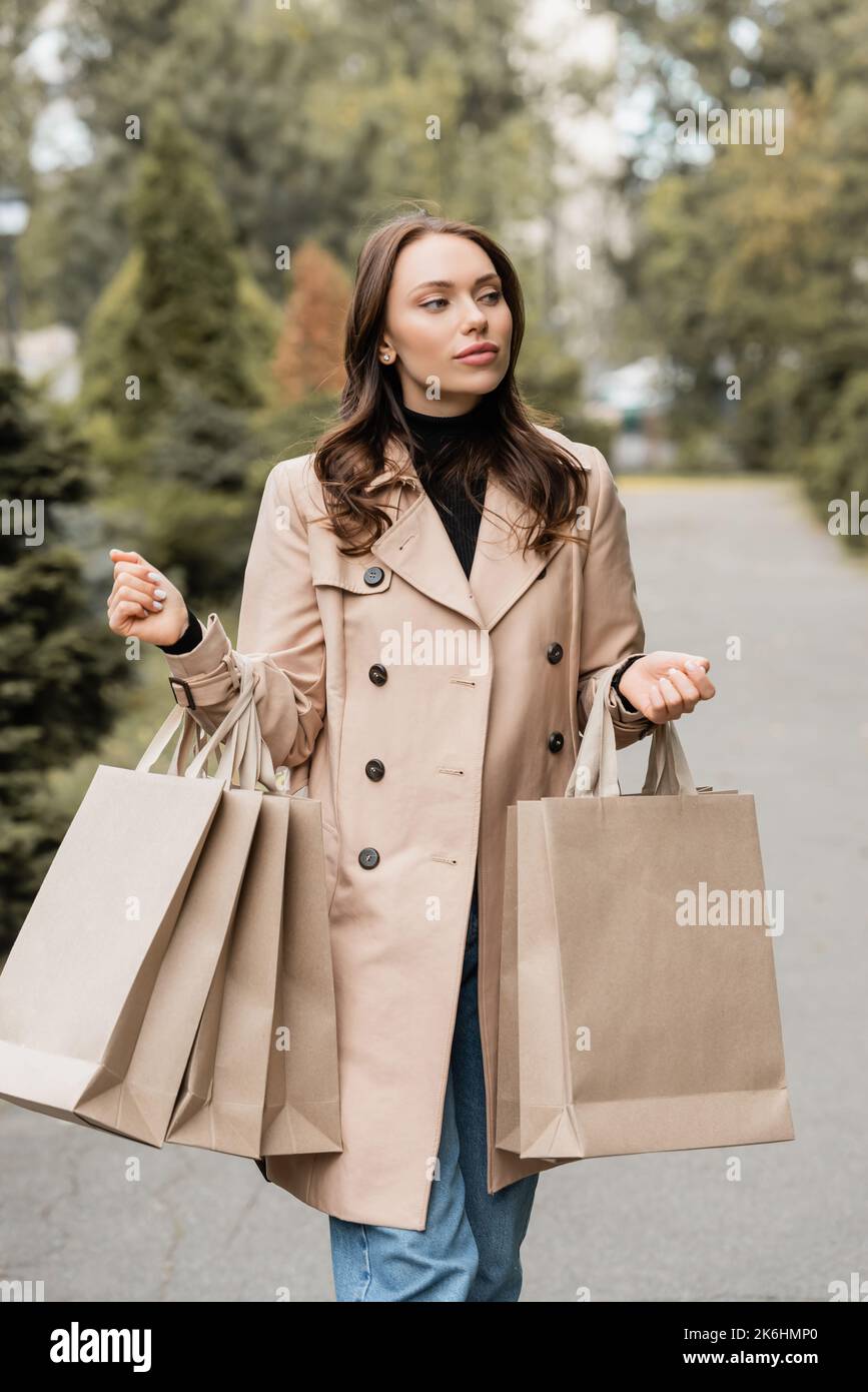Hübsche junge Frau im Trenchcoat, die Einkaufstaschen im herbstlichen Park hält, Stockbild Stockfoto