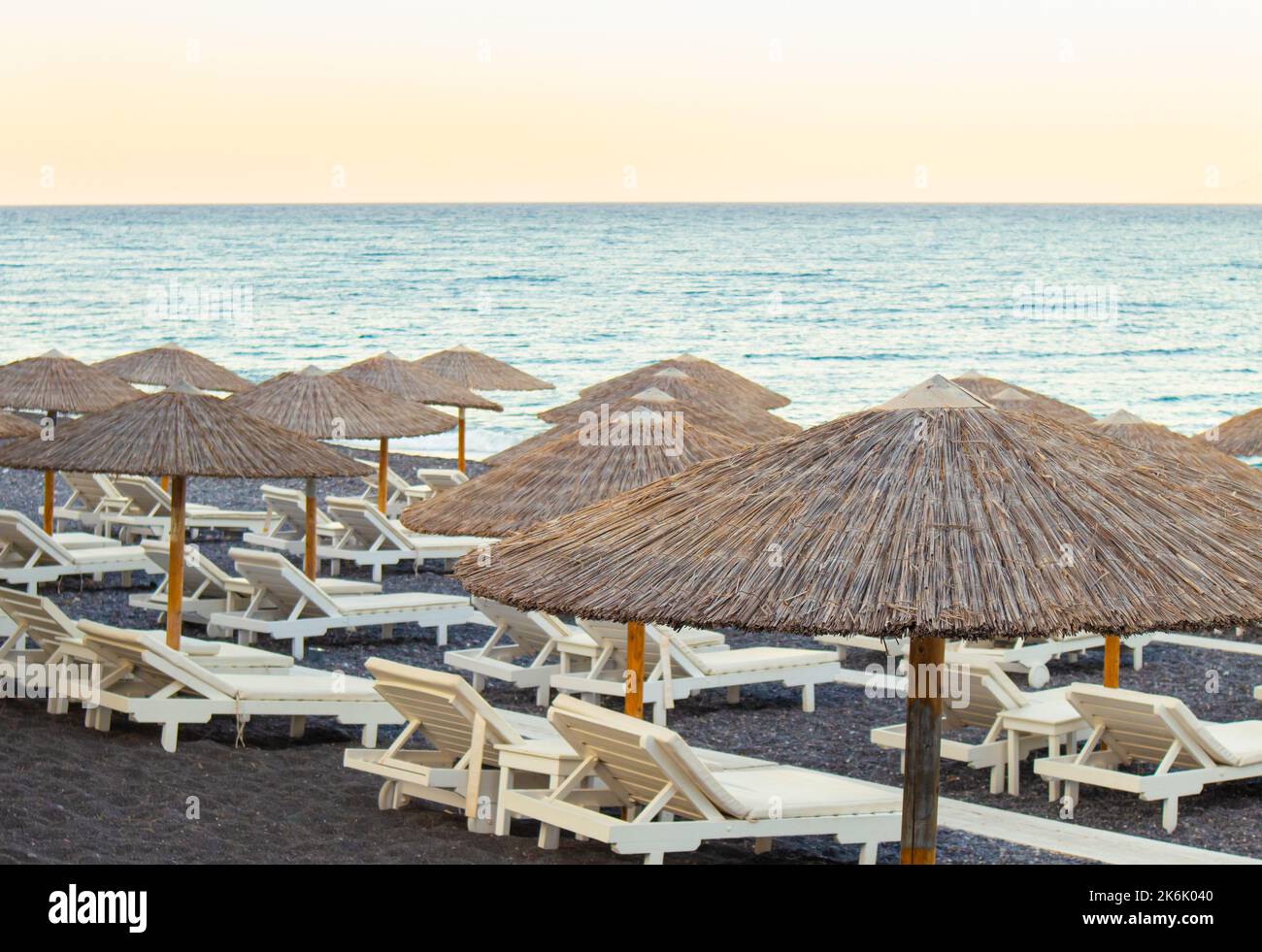 Weiße Sonnenstühle am schwarzen Sandstrand mit natürlichen Schilfschirmen bei Sonnenuntergang. Urlaub in Griechenland Konzept, viel Platz für Kopien. Stockfoto