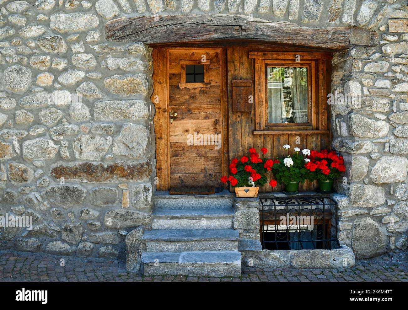Außenansicht einer typischen Steinhütte mit hölzerner Eingangstür und blühenden Geranien im Sommer, Courmayeur, Aostatal, Italien Stockfoto