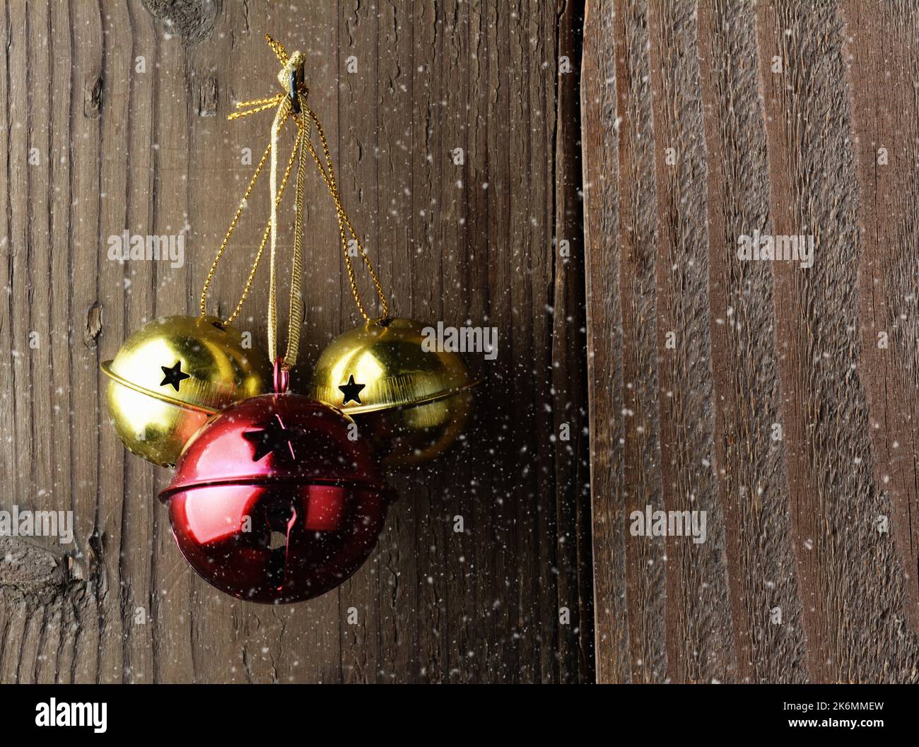 Rote und goldene Jingle-Glocken hängen an einer rustikalen Holzwand mit Schnee-Effekt. Stockfoto