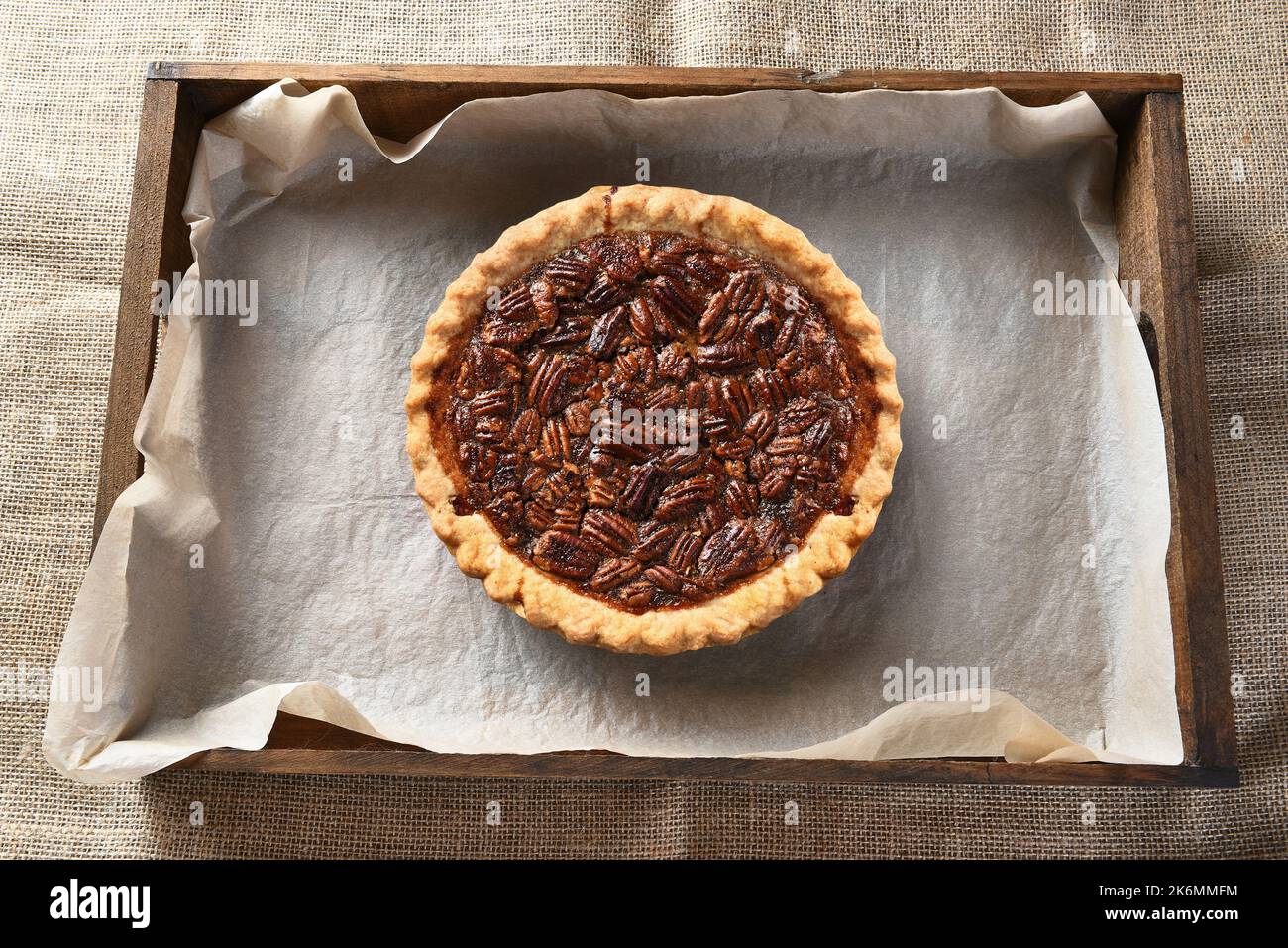 Ansicht eines Pecan-Kuchens in einer Holzkiste mit Pergamentpapier von oben. Der frisch gebackene Kuchen kühlt auf einem Tisch mit einem Lappen-Tischtuch ab. Stockfoto