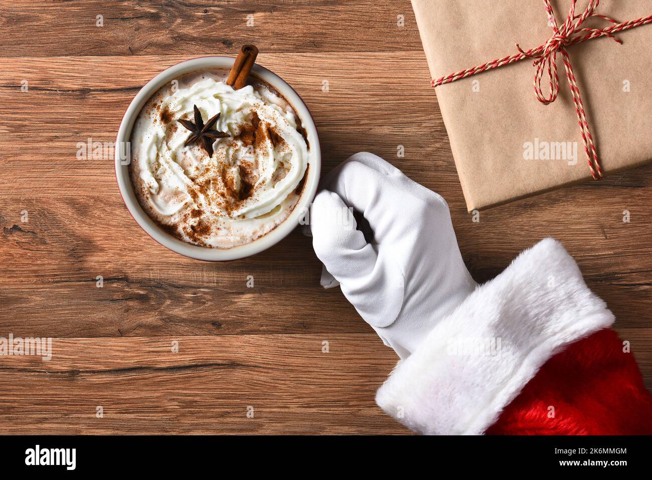 Nahaufnahme des Weihnachtsmannes mit einem Becher heißen Kakaos auf einem Holztisch und einem schlichten, verpackten Geschenk. Stockfoto