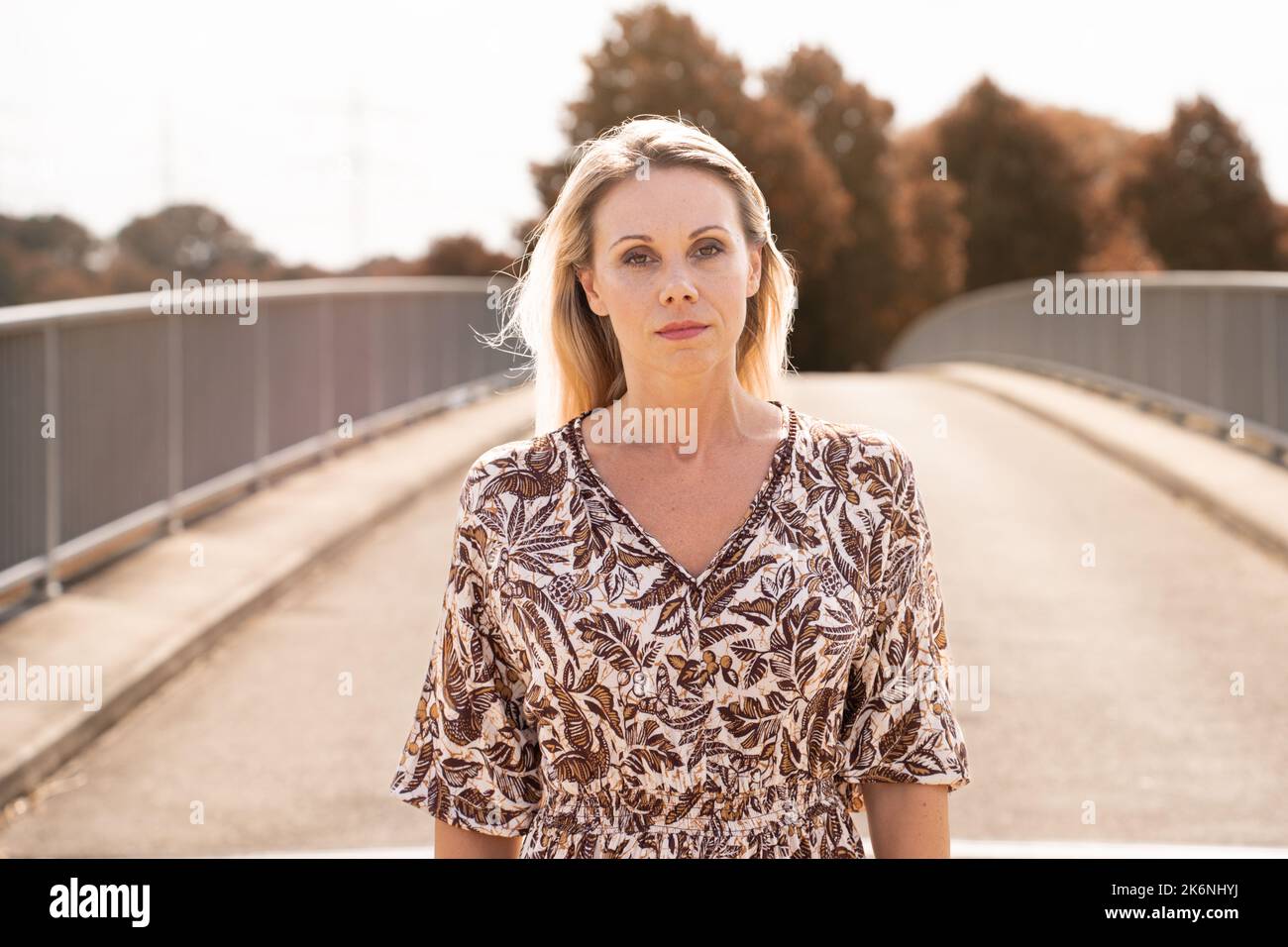 Frontalporträt einer blonden Frau mittleren Alters mit einem fokussierten Blick auf die Kamera vor einem herbstlichen Hintergrund, der auf einer Brücke läuft Stockfoto
