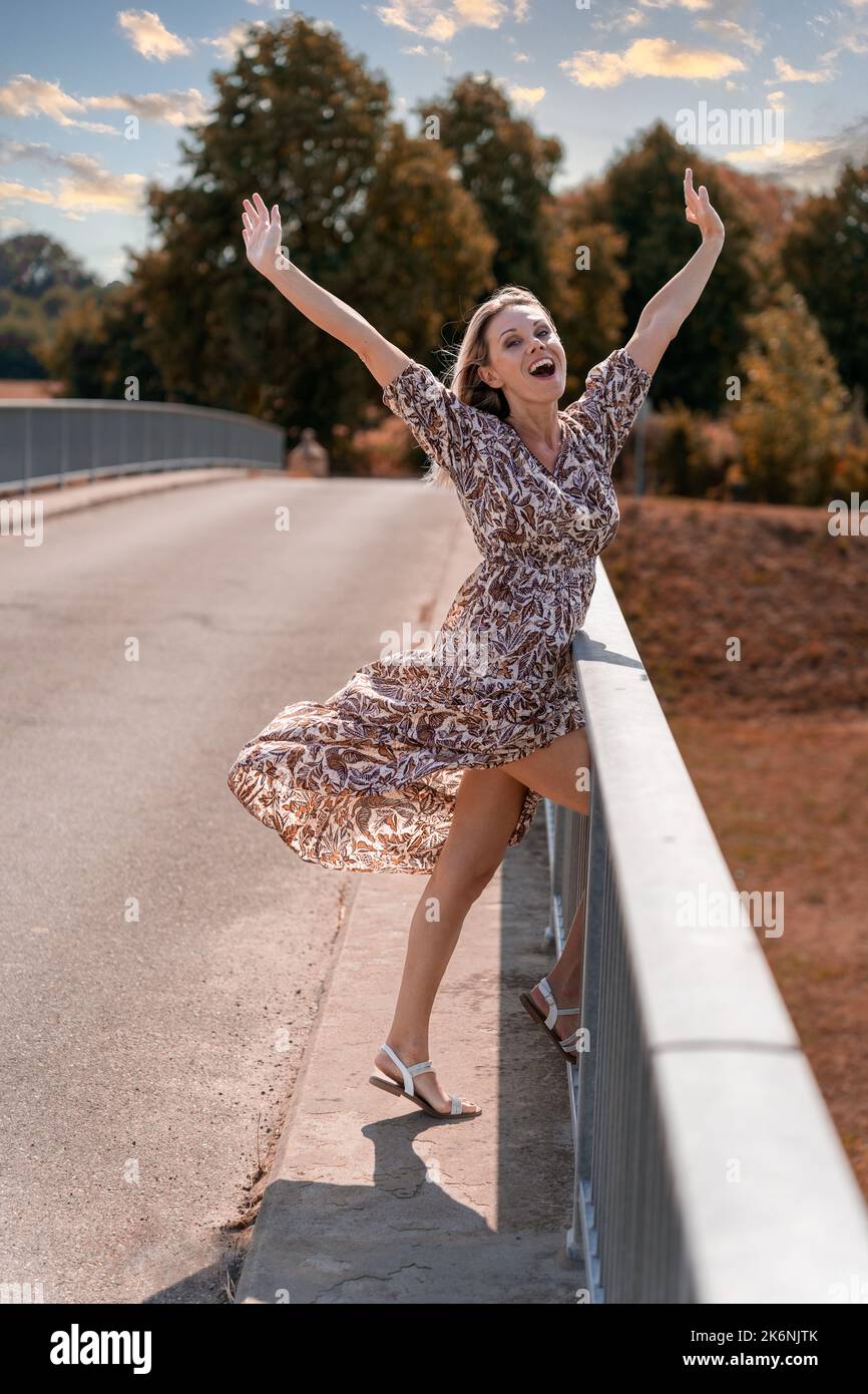 Attraktive, enthusiastisch jubelende blonde Frau mittleren Alters, die beide Arme in den Himmel ragte, um sich über den Erfolg auf einer Brücke in herbstlichen Farben zu freuen Stockfoto