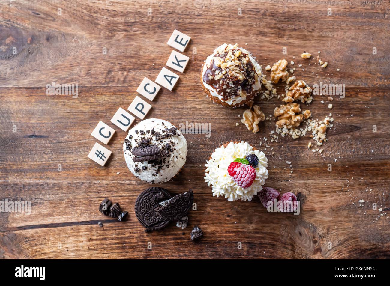Drei leckere Cupcakes auf einem Holztisch. Weiße Creme, Schokolade und Himbeere, mit Nuss überzogen. Das Wort Cupcake aus Holzblöcken. Stockfoto