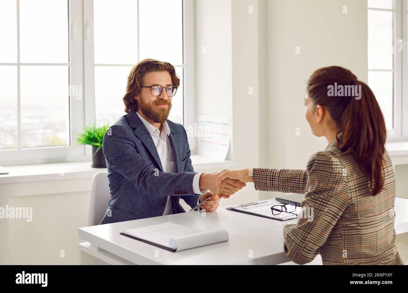 Zufriedener Agent und Kunde, der am Schreibtisch sitzt, ein Geschäft abmacht, lächelt und die Hände schüttelt Stockfoto