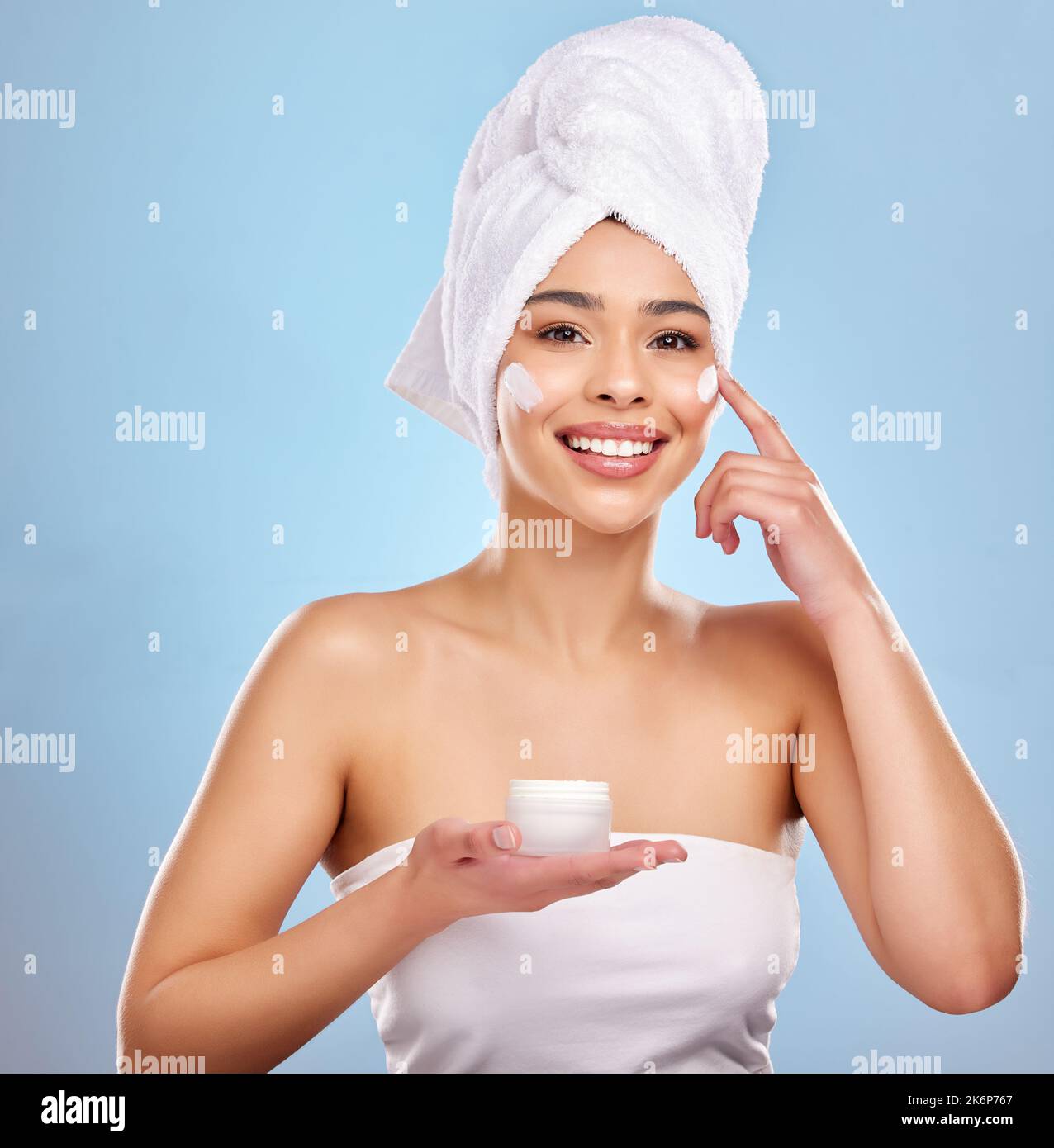 Heute leuchten, jeden Tag leuchten. Studioporträt einer attraktiven jungen Frau, die auf ihrem Gesicht vor blauem Hintergrund Feuchtigkeitscreme auftragt. Stockfoto