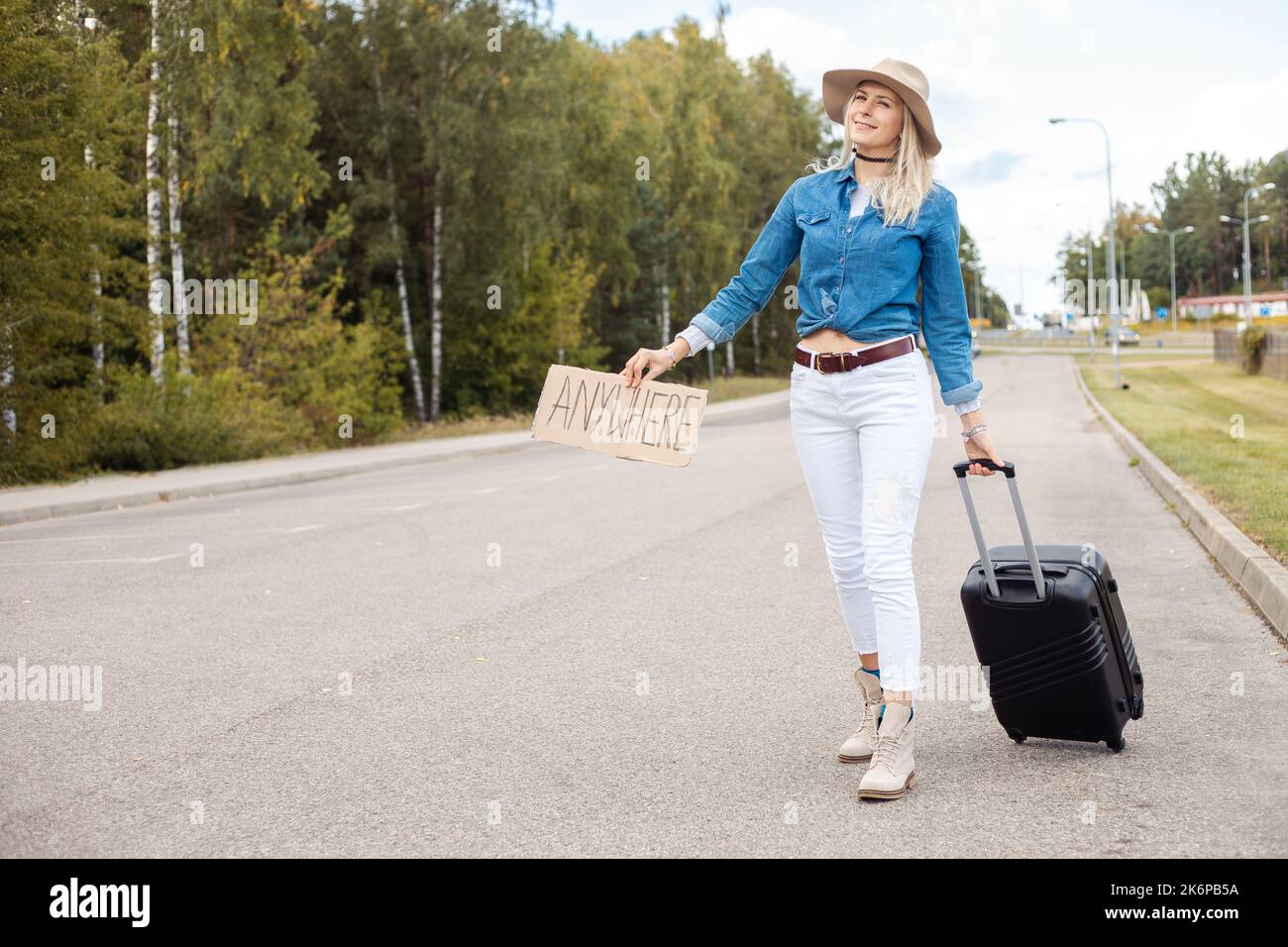 Fröhliche Frau wartet auf leerer Straße mit Pappkarton und Koffer. Lady im Hut- und Denim-Outfit entfliehen Sie per Auto-Stop-to-Go aus der Stadt Stockfoto
