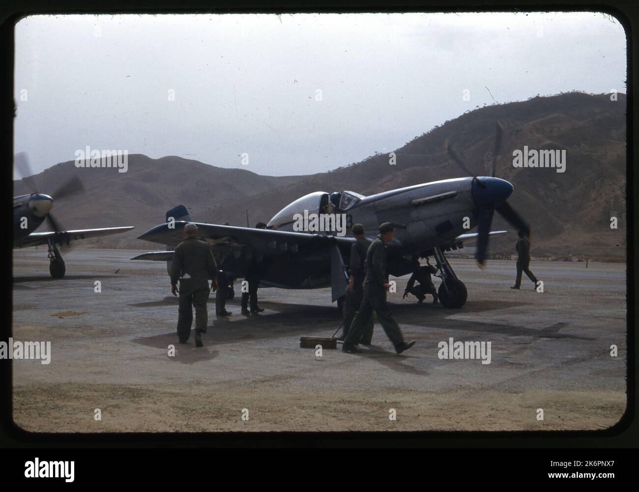 Ein Viertel von rechts Vorderansicht eines nordamerikanischen F-51D Mustang, der irgendwo in Korea auf dem Hardstand geparkt war. Mehrere Besatzungsmacht erscheinen um das Flugzeug herum, um die letzten Vorbereitungen für den Start zu treffen, und ein Pilot ist im Cockpit. Die Nase und der linke Flügel eines anderen F-51D Mustang sind sichtbar, die hinter dem Flugzeug geparkt sind. Stockfoto