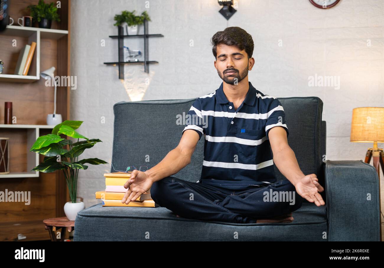 Ein junger indischer Junge, der vor dem Lesen mit geschlossenen Augen meditiert, während er zu Hause auf dem Sofa sitzt - Konzept der Stressbewältigung, des Wissens und der Entspannung. Stockfoto