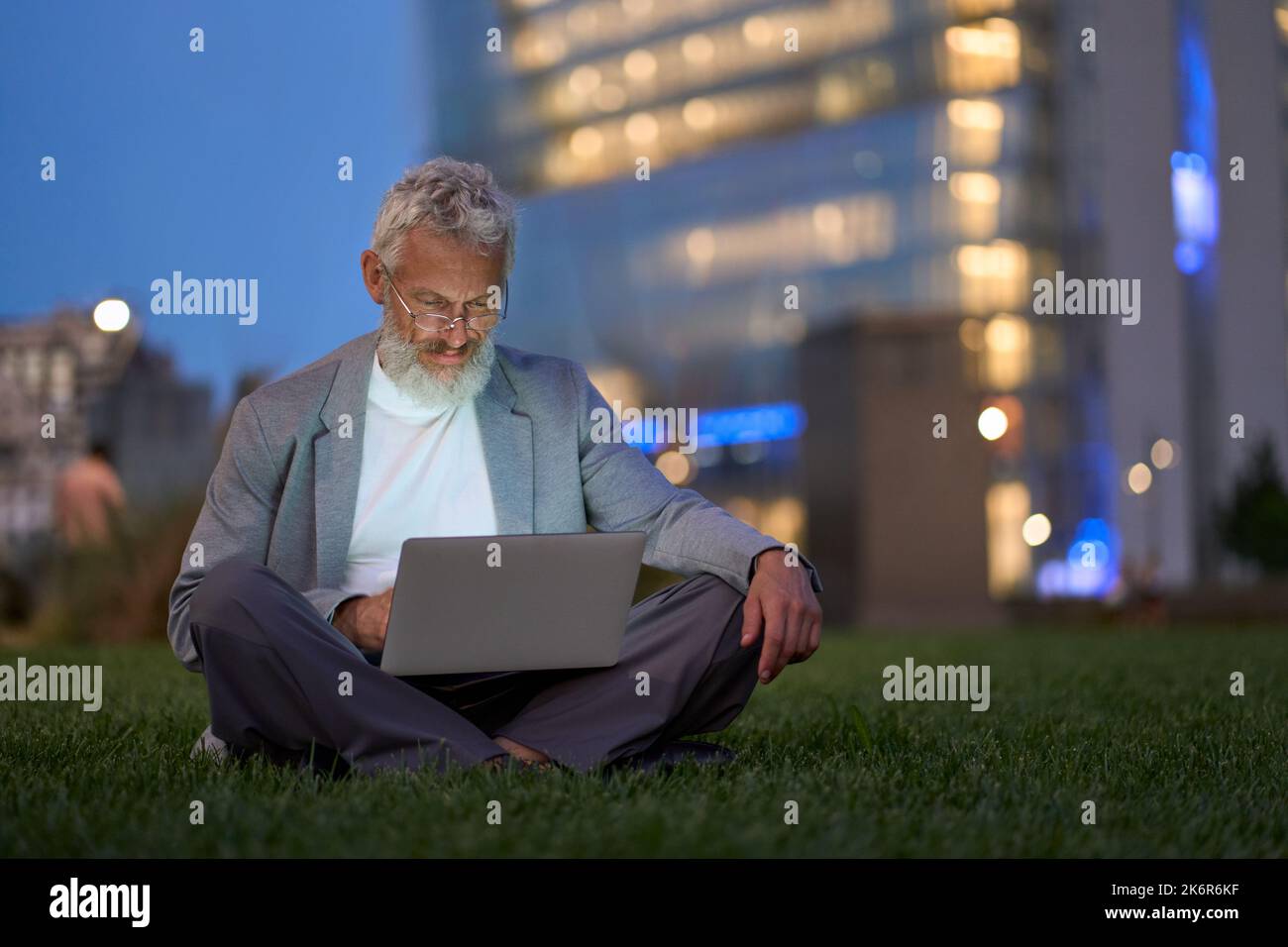 Ein professioneller älterer Geschäftsmann, der im nächtlichen Stadtpark einen Laptop benutzt. Stockfoto
