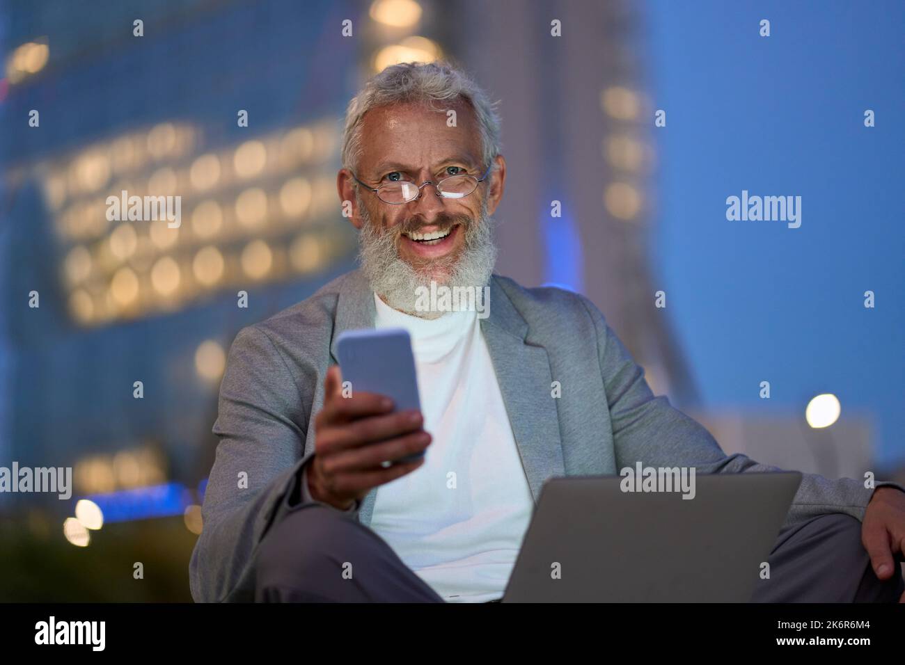 Glücklicher alter Geschäftsmann, der nachts im Stadtpark vor dem Büro sitzt und telefoniert. Stockfoto