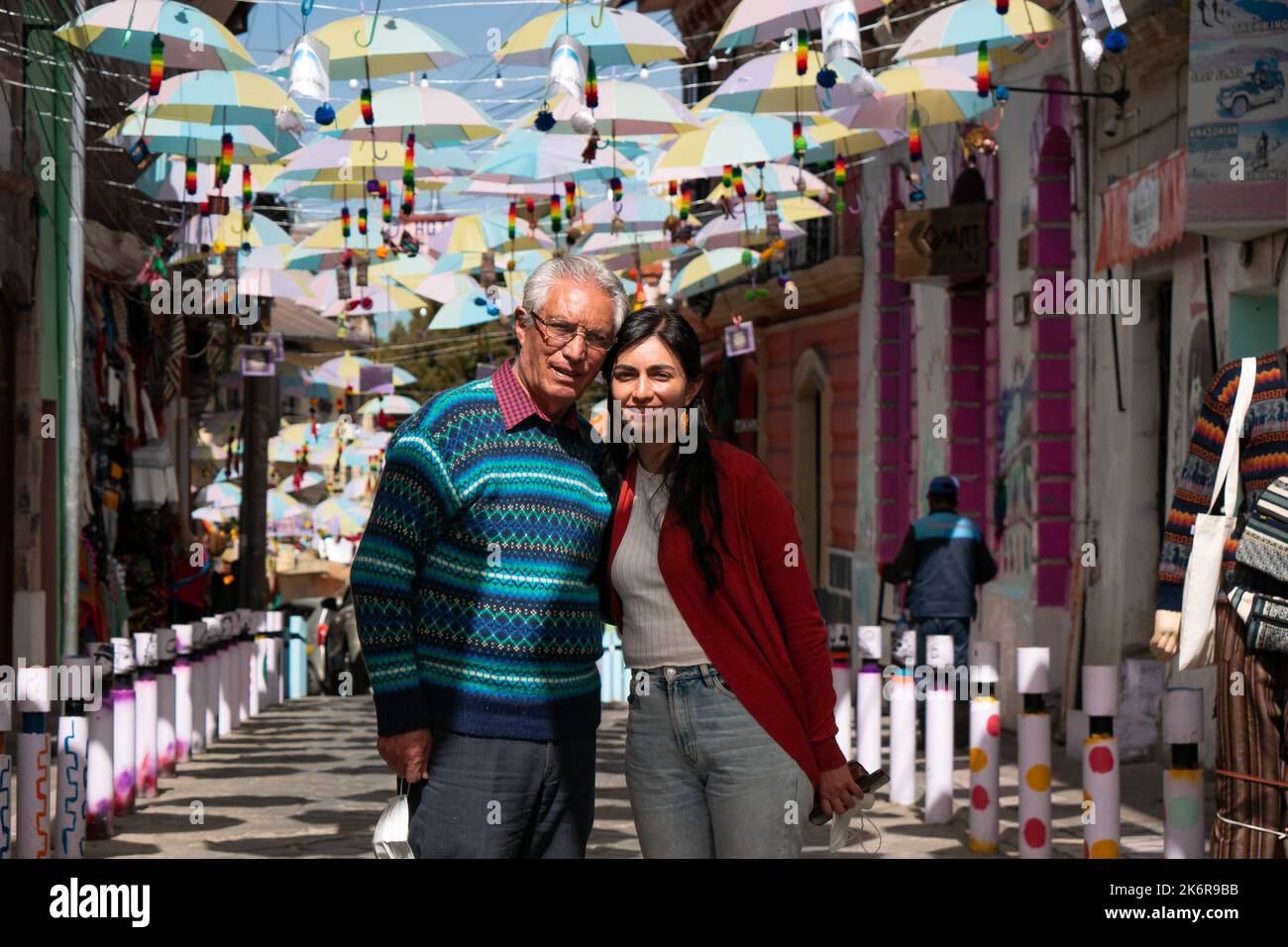 Die junge schwarzhaarige bolivianische Frau und ein älterer grauhaariger peruanischer Mann, der eine Brille trägt, posieren auf einer Straße, die mit Regenschirmen am Himmel gesäumt ist Stockfoto
